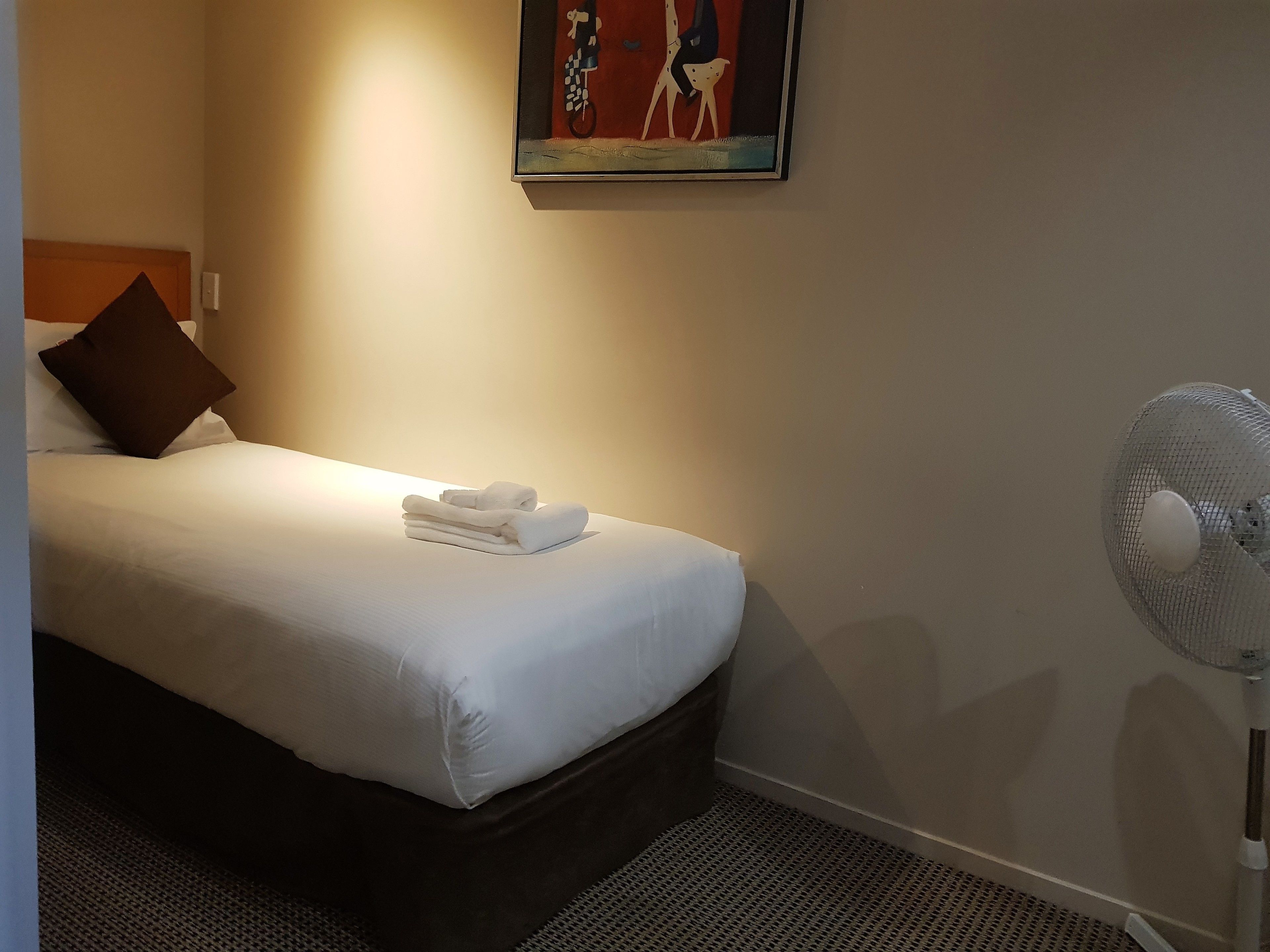 A neatly made bed with white linens and a folded towel, a standing fan, and a painting on the wall.