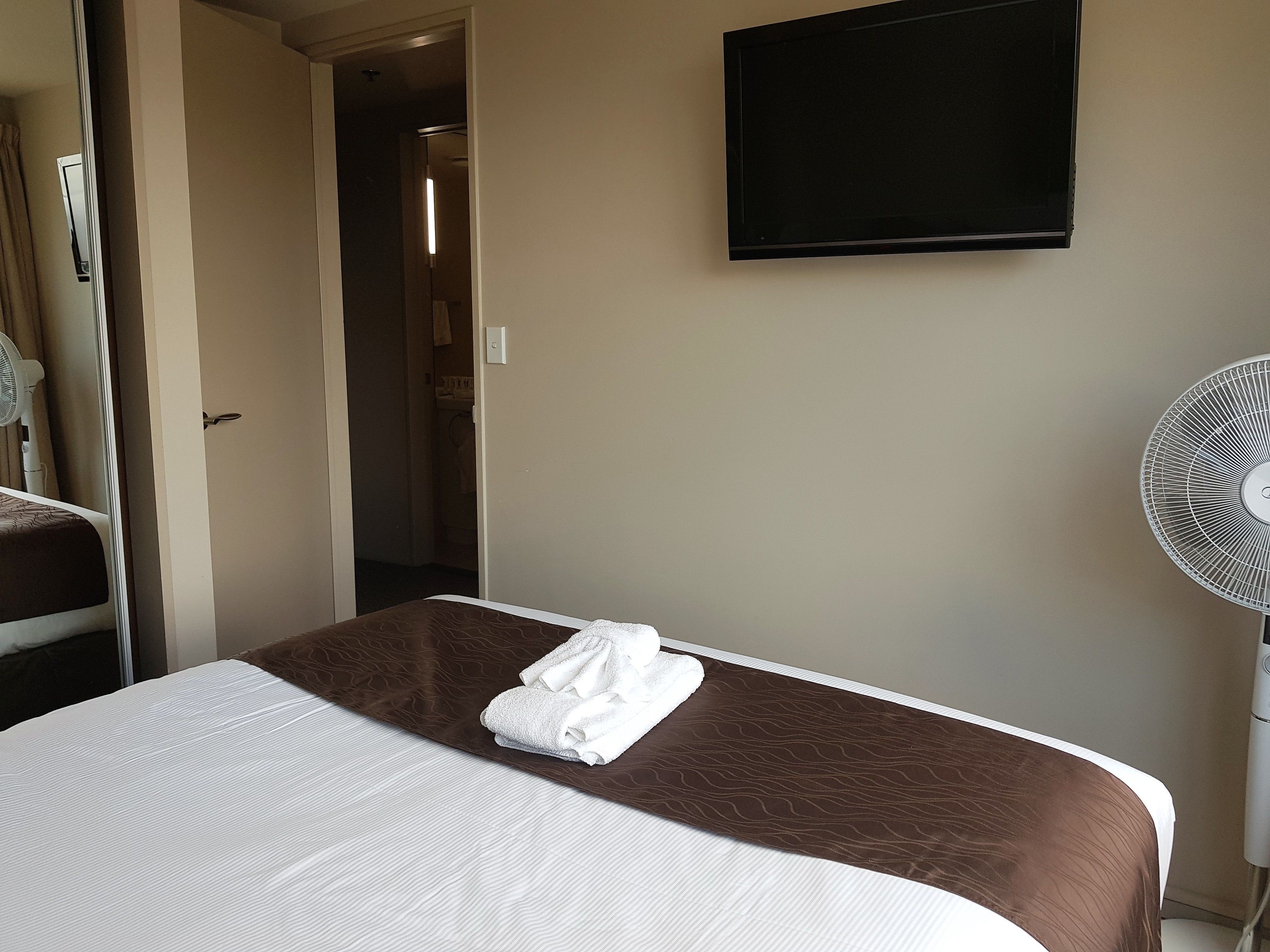 A hotel room with a bed, a flat-screen TV mounted on the wall, a standing fan, and a door leading to a bathroom.