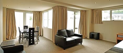 A modern hotel room with a dining area, a sofa, and large windows with curtains.