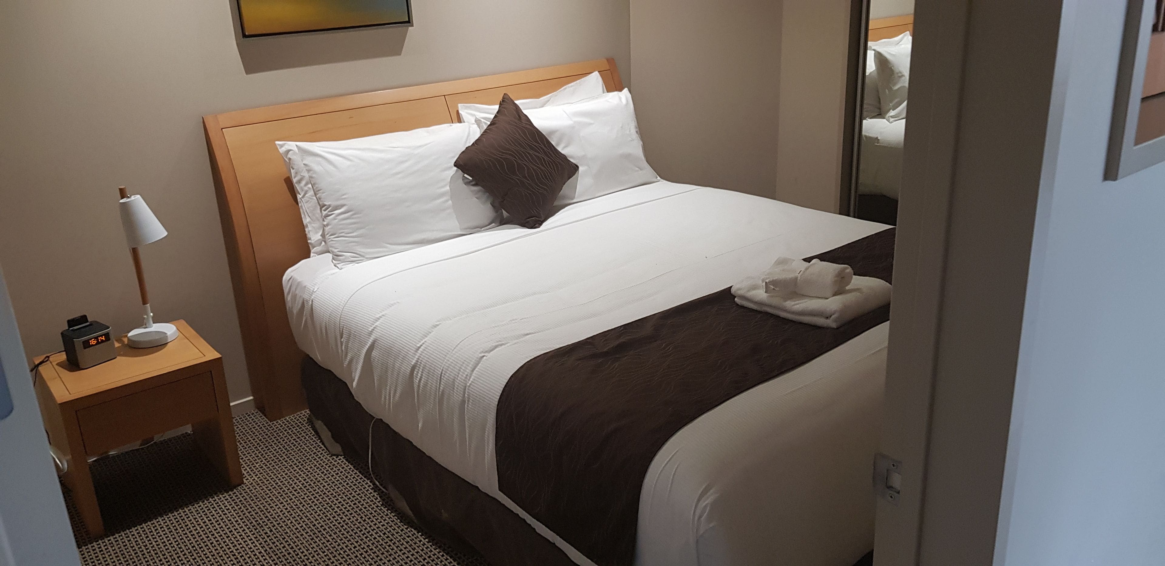 Uma cama bem arrumada com roupa de cama branca e marrom, uma luminária de cabeceira e um criado-mudo com uma lâmpada e um dispositivo.