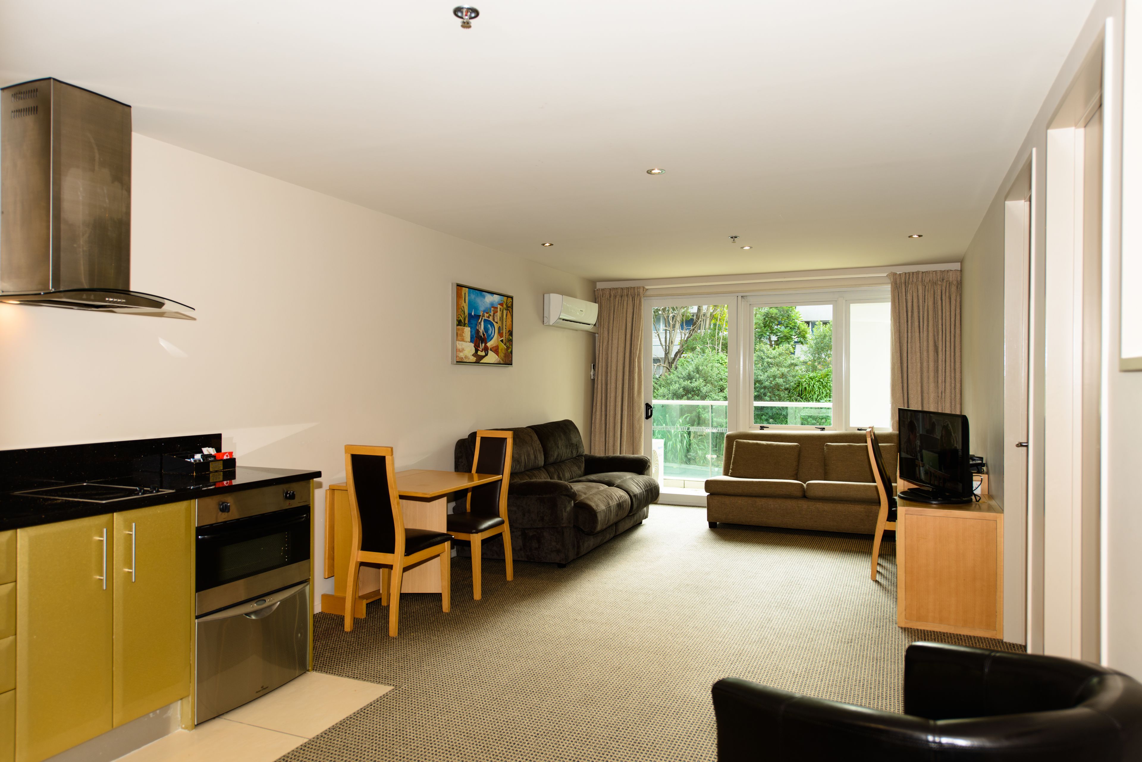 A modern kitchen with stainless steel appliances, a dining area with a table and chairs, a living room with a sofa and a television, and a large sliding glass door leading to a balcony.