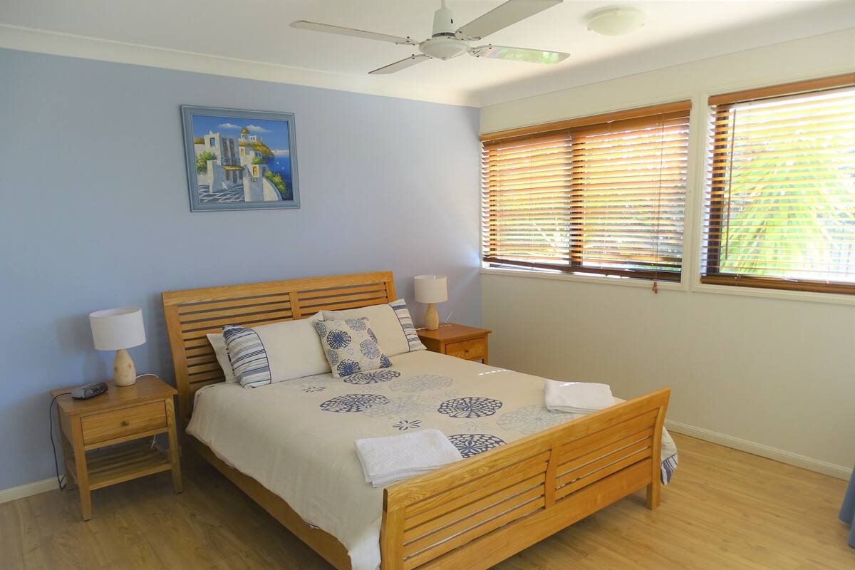 A bedroom with a wooden bed, two bedside tables, a ceiling fan, and a window with blinds.