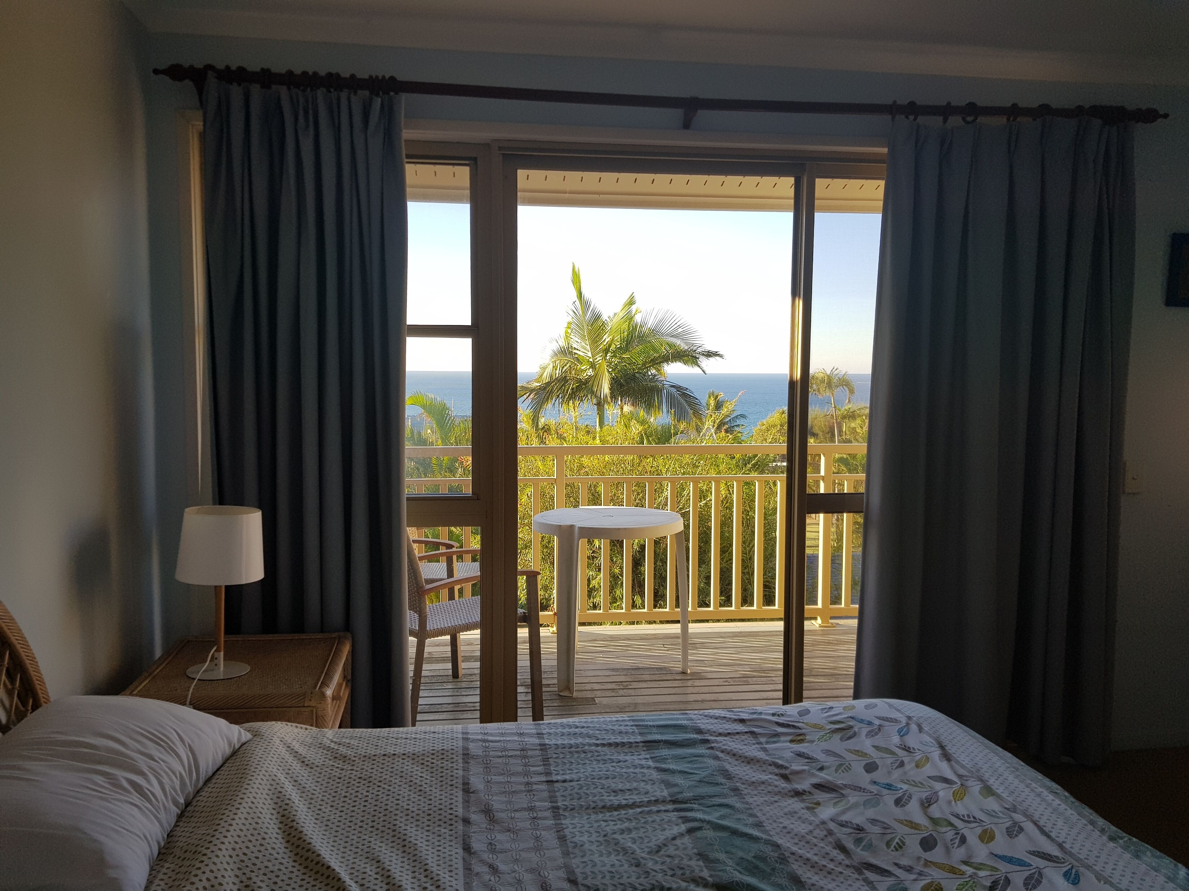 A bedroom with a bed, a nightstand, a lamp, a view of the ocean, and a balcony with a table and chair.