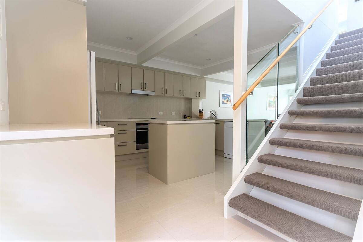 A modern kitchen with a central island, a built-in oven, and a glass staircase leading to a landing area.