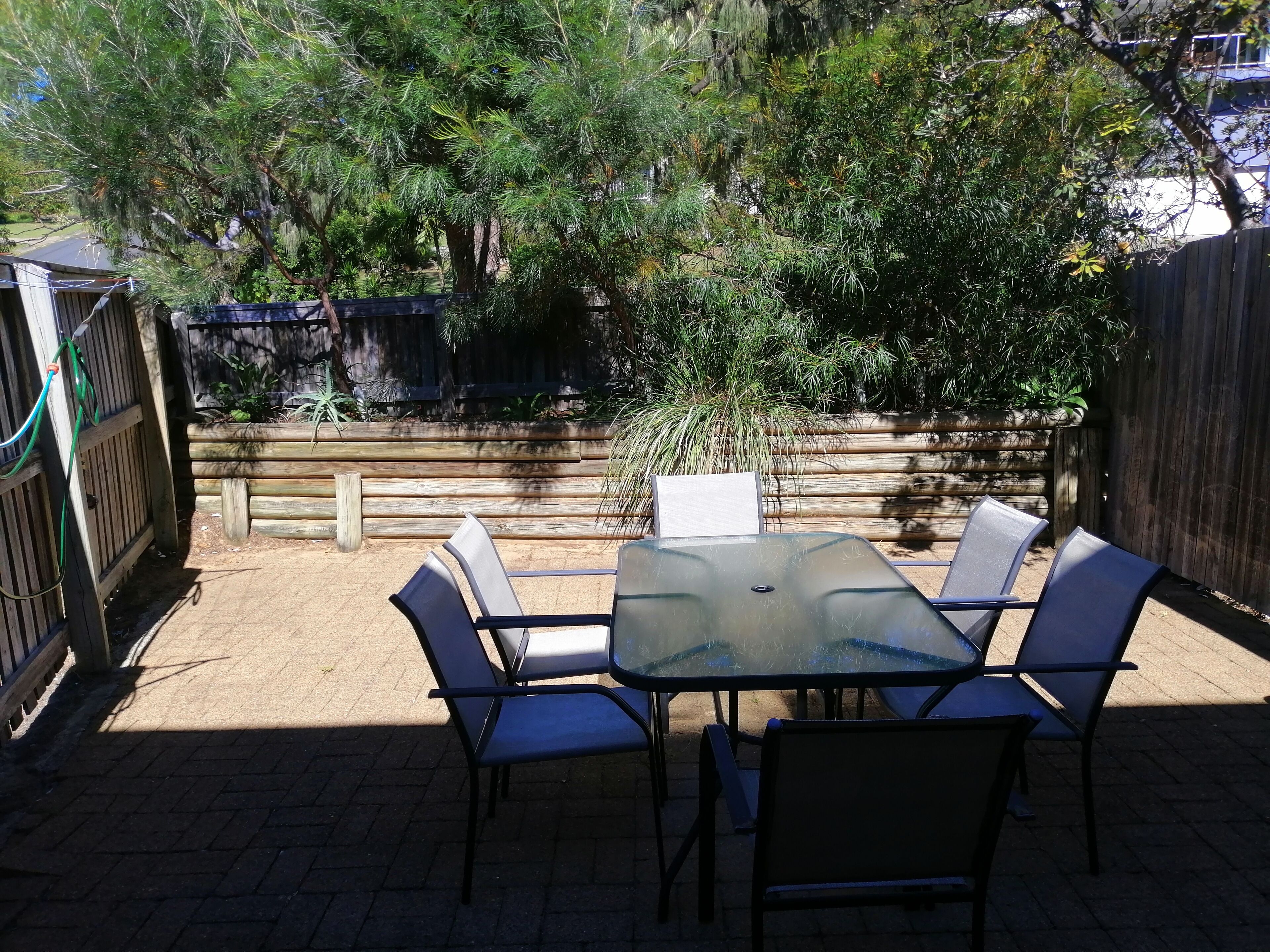 Une terrasse avec une table et des chaises en verre, entourée d’une clôture en bois et de verdure.