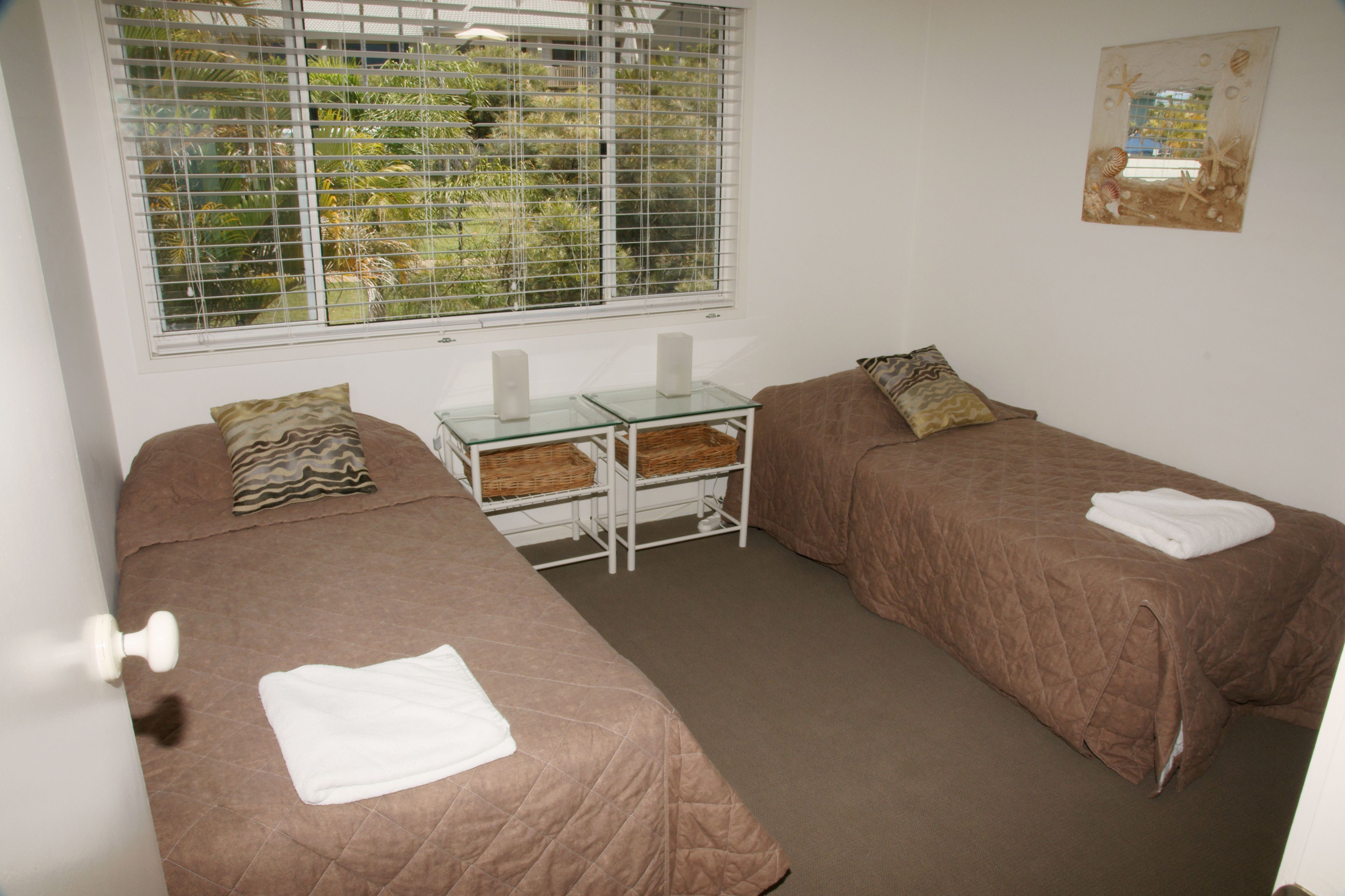 A room with two single beds, a small table with a glass top, and a window with blinds.