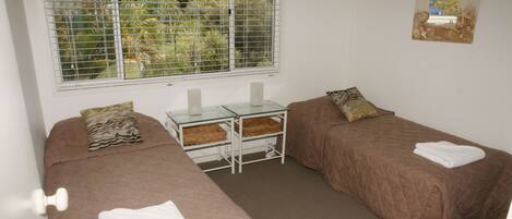 A room with two single beds, a small table with a glass top, and a window with blinds.