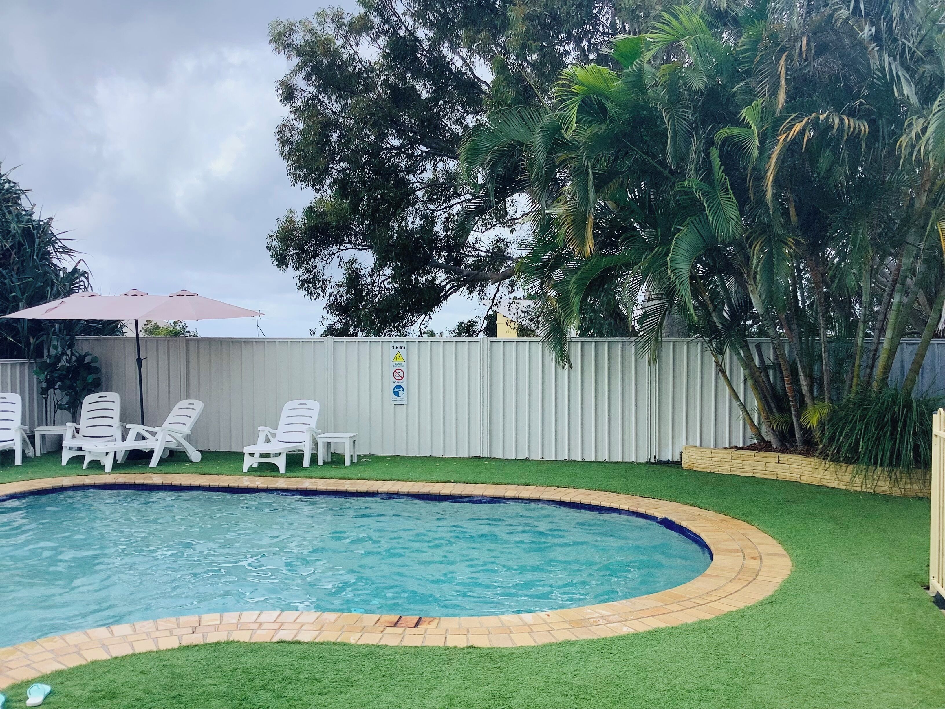 Une piscine entourée d’une pelouse, de chaises longues blanches et d’une clôture portant un panneau d’avertissement concernant la profondeur de l’eau.