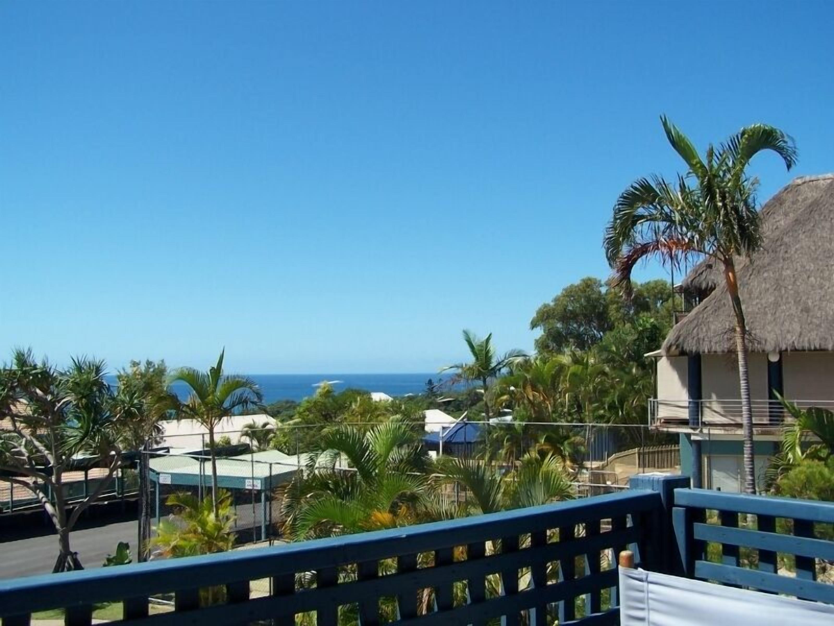 A view from a balcony overlooking a beach with palm trees and a thatched-roof building.