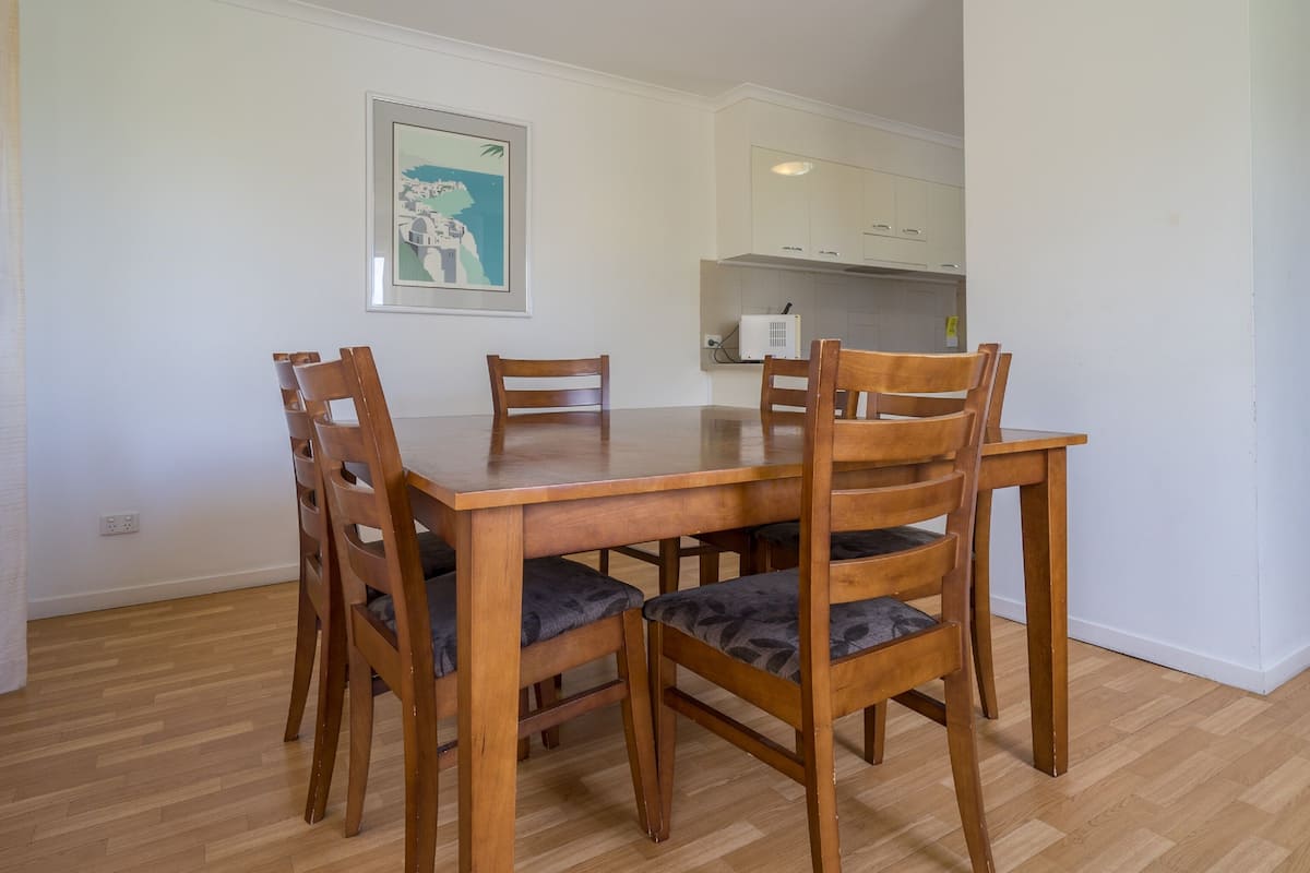 A dining area with a wooden table and chairs, a kitchen in the background, and a framed picture on the wall.