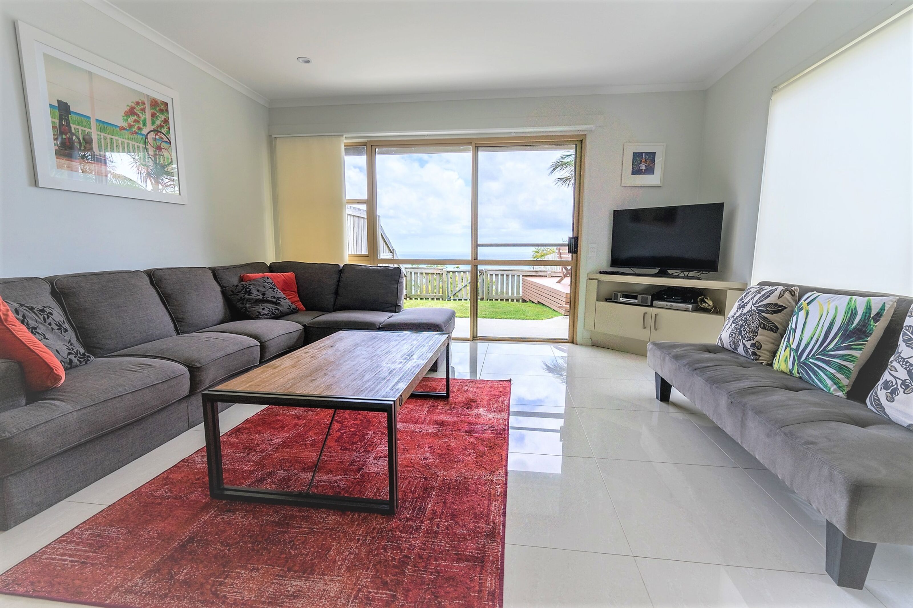 A modern living room with a grey sectional sofa, a wooden coffee table, and a red rug. There is a television on a stand and a balcony with a view of the ocean.