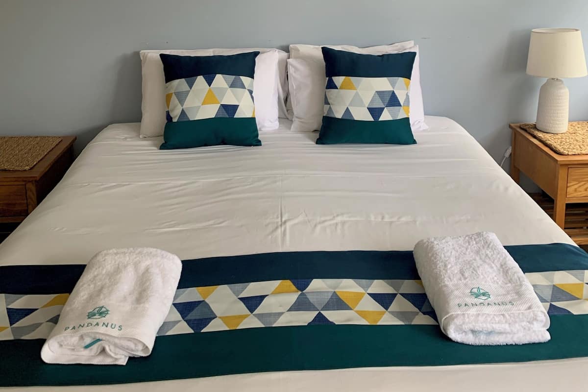 A neatly made bed with a geometric-patterned pillow, a framed painting of a coastal building, and a ceiling fan.