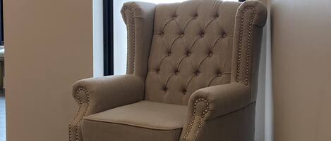 A tufted armchair with a high back and decorative beading, positioned in a hallway with a tiled floor and a large window with blinds.