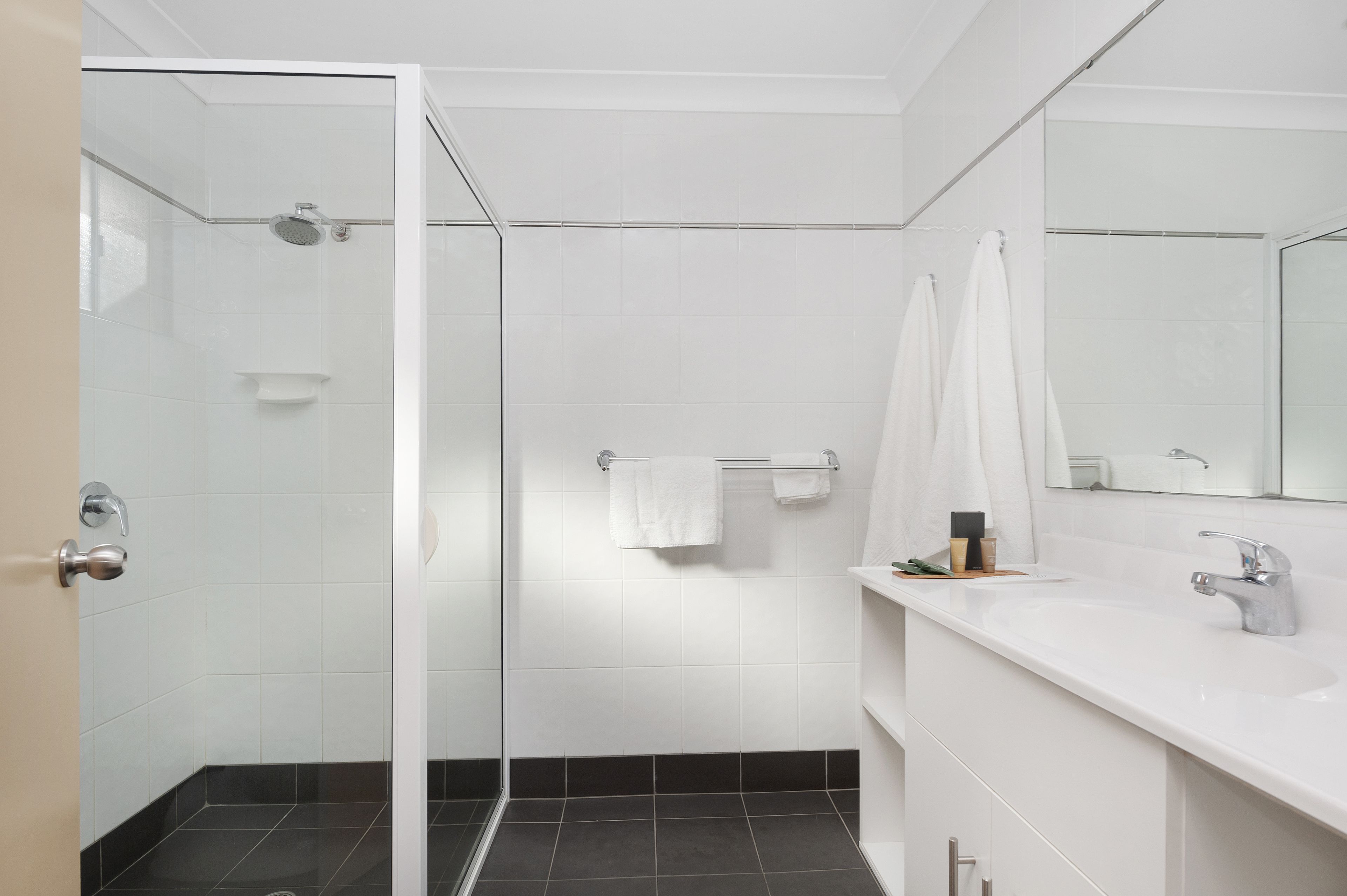 A modern bathroom with a glass-enclosed shower, white tiled walls, a large mirror, and a white sink.