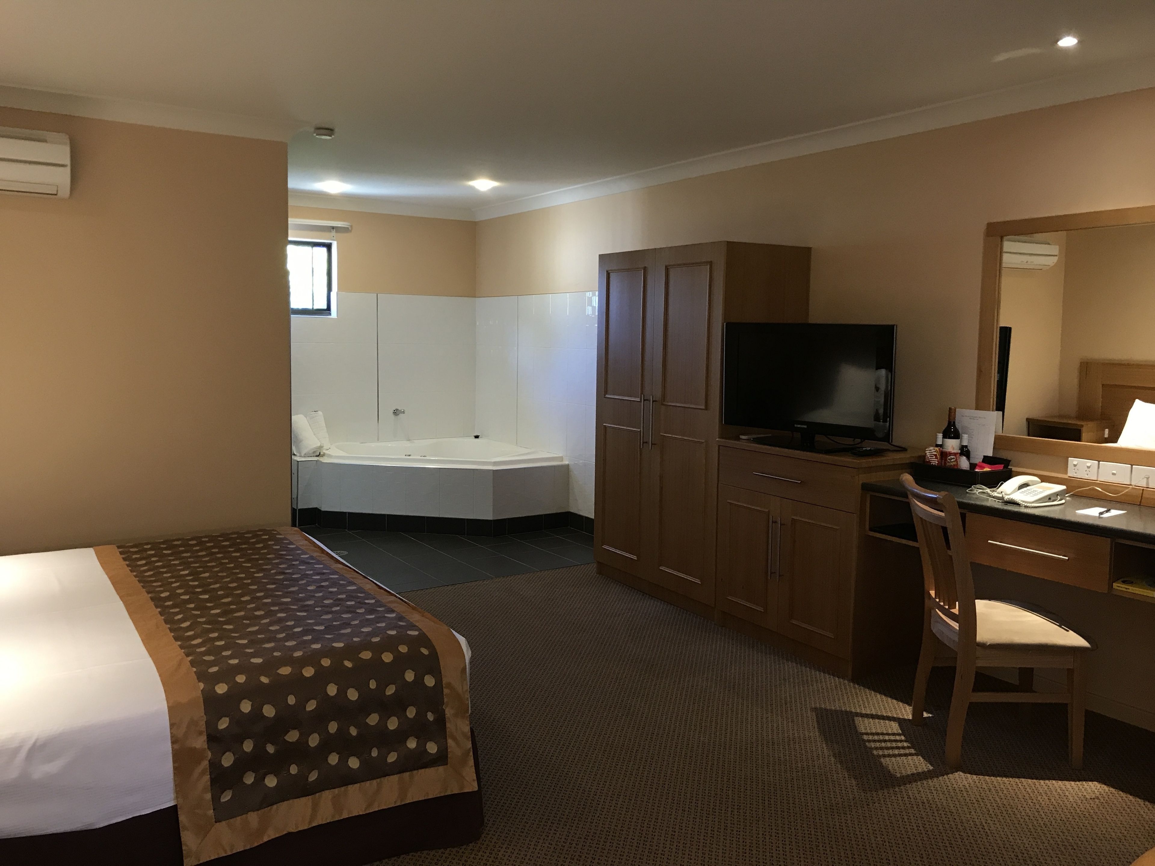Quarto de hotel com cama, escrivaninha com cadeira, televisão, minibar e banheiro com banheira e chuveiro.