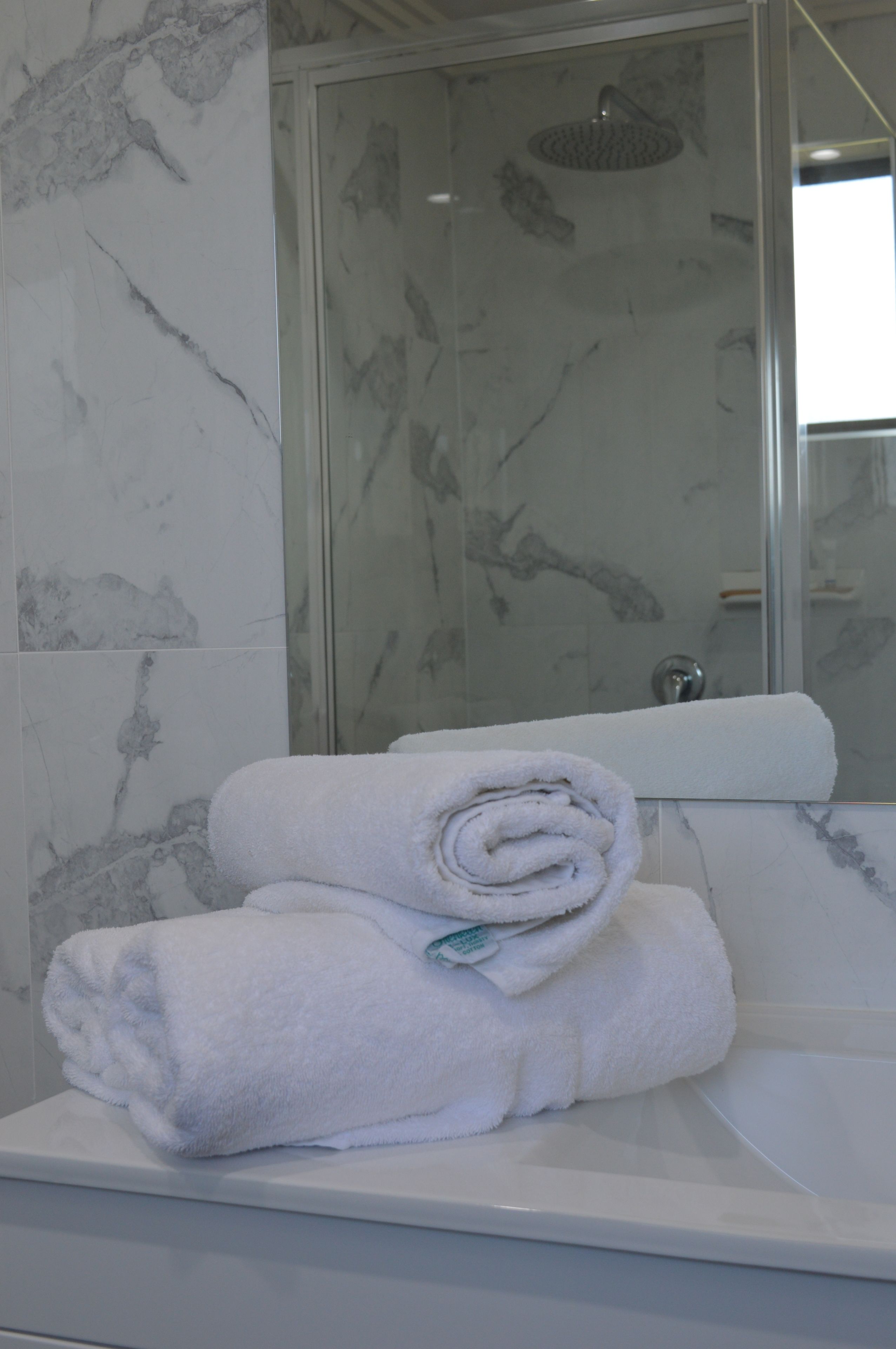 A bathroom with marble walls, a glass shower enclosure, and a stack of white towels on a white sink.