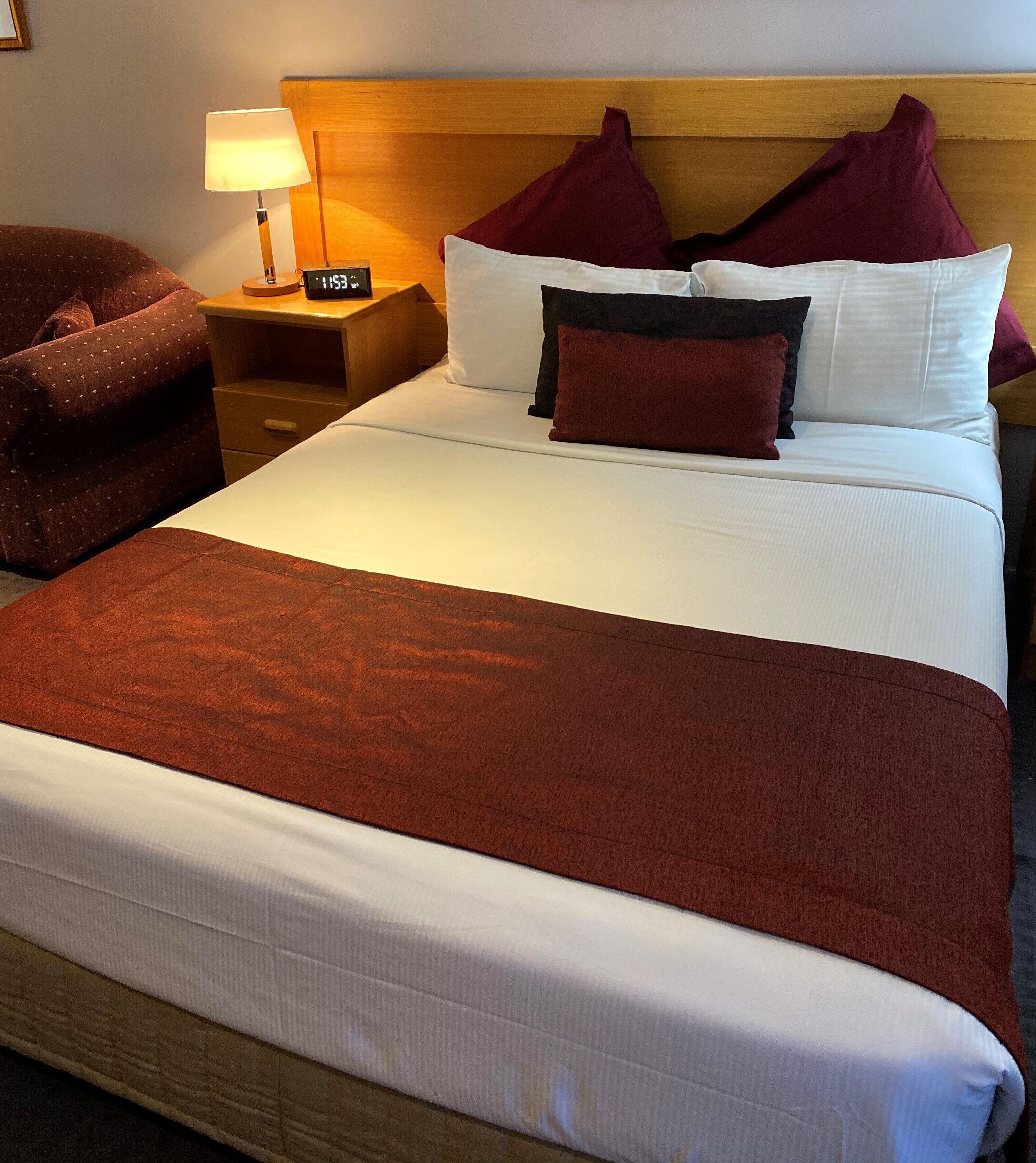 Una cama bien tendida con tablero de madera, una mesita de noche con lámpara y un sillón marrón.