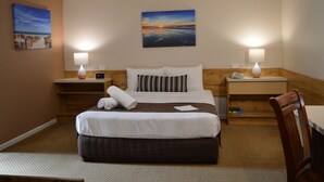 Habitación de hotel con cama, dos mesitas de noche con lámparas, cabecera de madera, un cuadro en la pared y vistas del atardecer.