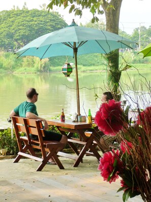 Een houten tafel met stoelen onder een blauwe parasol, met uitzicht op een kalm meer en begroeiing op de achtergrond.