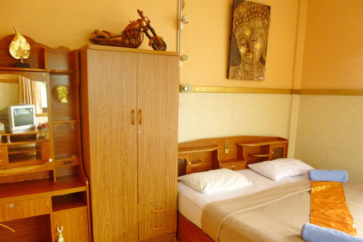 Een hotelkamer met een houten garderobe, een bed met wit beddengoed, een klein bureau met een televisie, en een decoratief Boeddhabeeld aan de muur.
