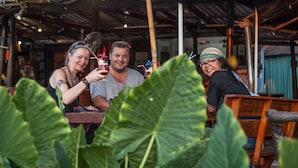 Een groep mensen zit aan een houten tafel in een rustieke binnenruimte, met grote groene bladeren op de voorgrond.