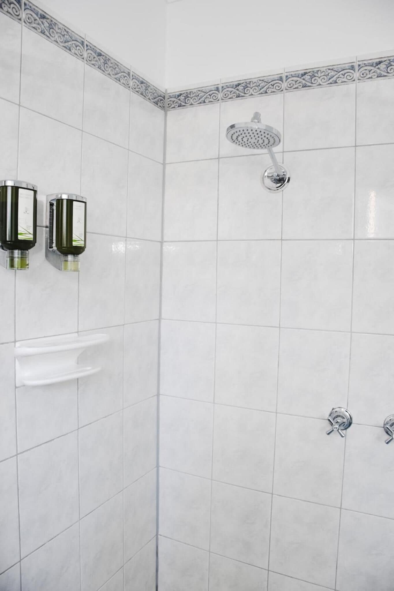 A shower area with white tiles, a showerhead, and two hand soap dispensers.