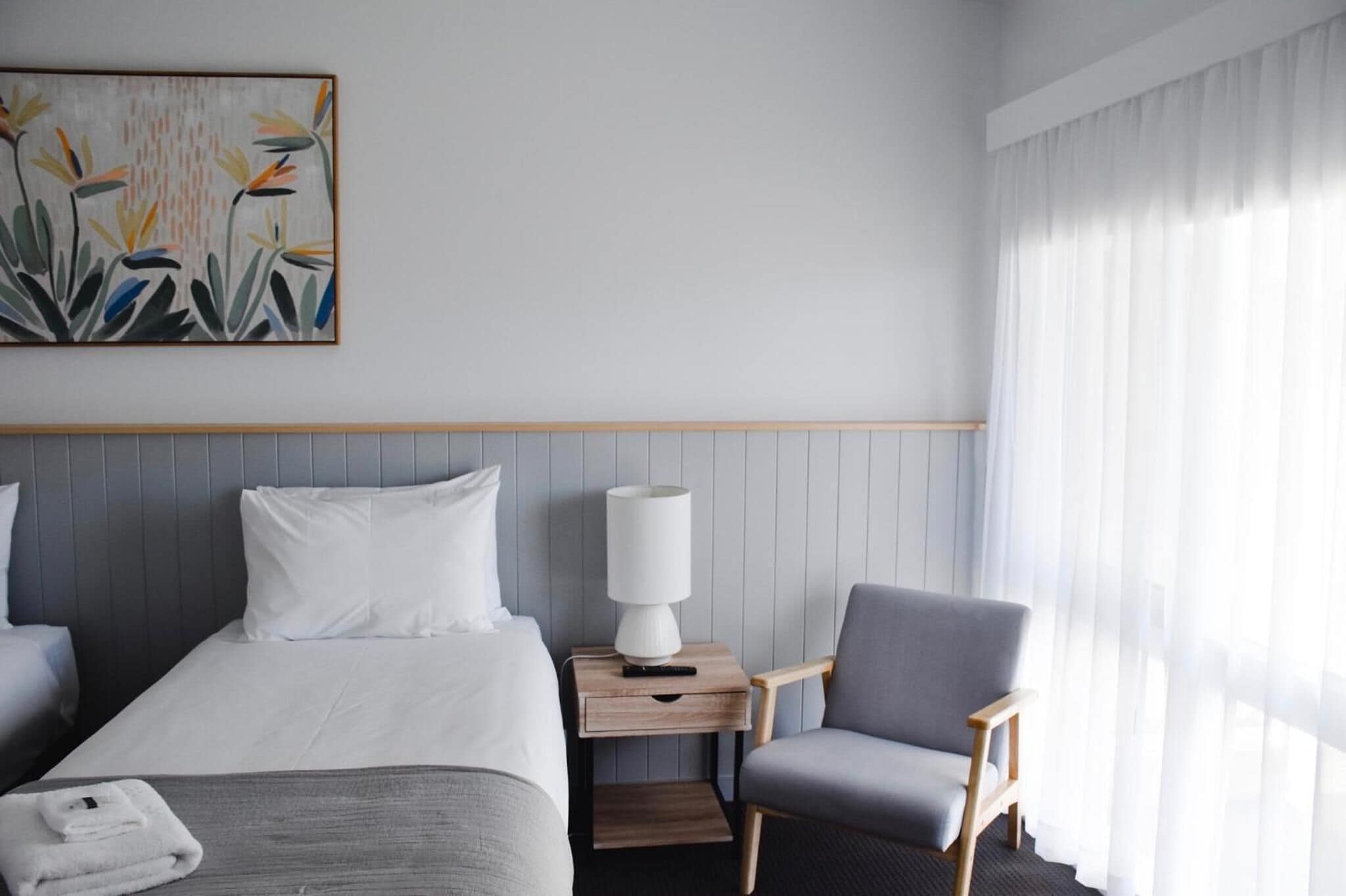 A hotel room with a bed, a chair, a nightstand with a lamp, and a painting on the wall.