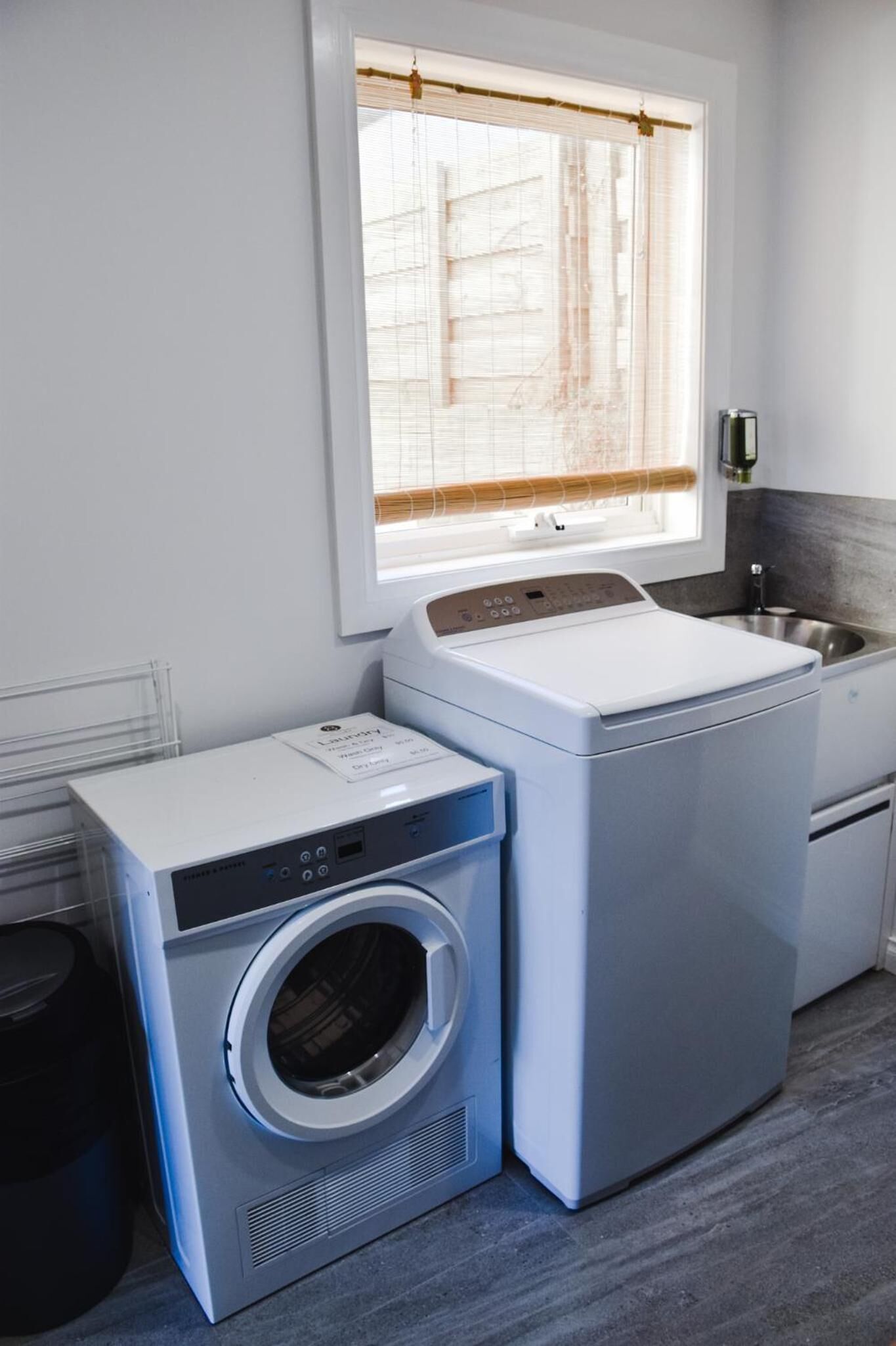 A compact laundry area with a front-loading washer and dryer, a small sink, and a window with blinds.