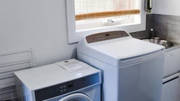 A compact laundry area with a front-loading washer and dryer, a small sink, and a window with blinds.