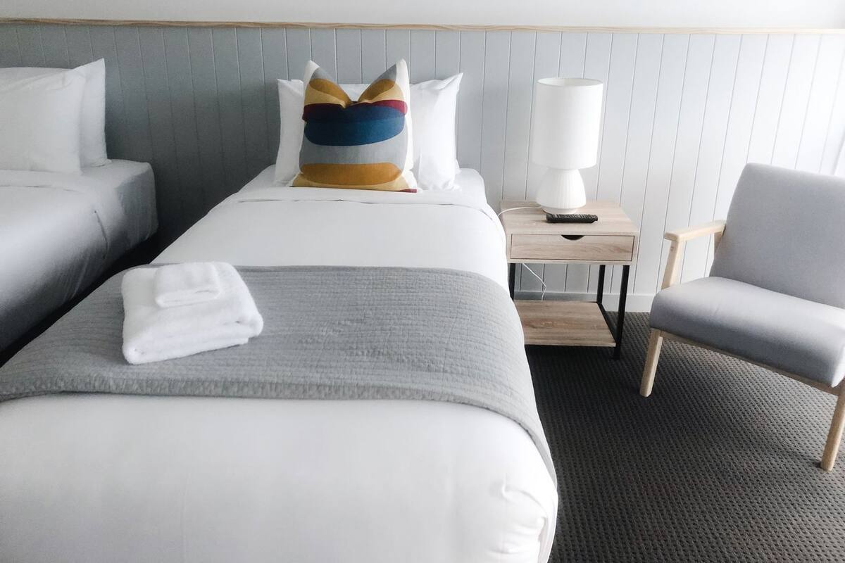 A hotel room with two beds, a bedside table with a lamp, a chair, and a window with blinds.