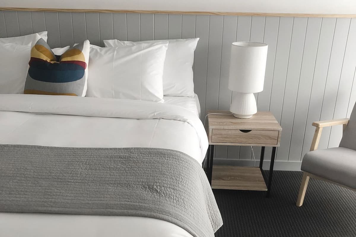 A neatly made bed with white bedding, a grey throw blanket, and colorful pillows. There is a bedside table with a lamp and a chair.