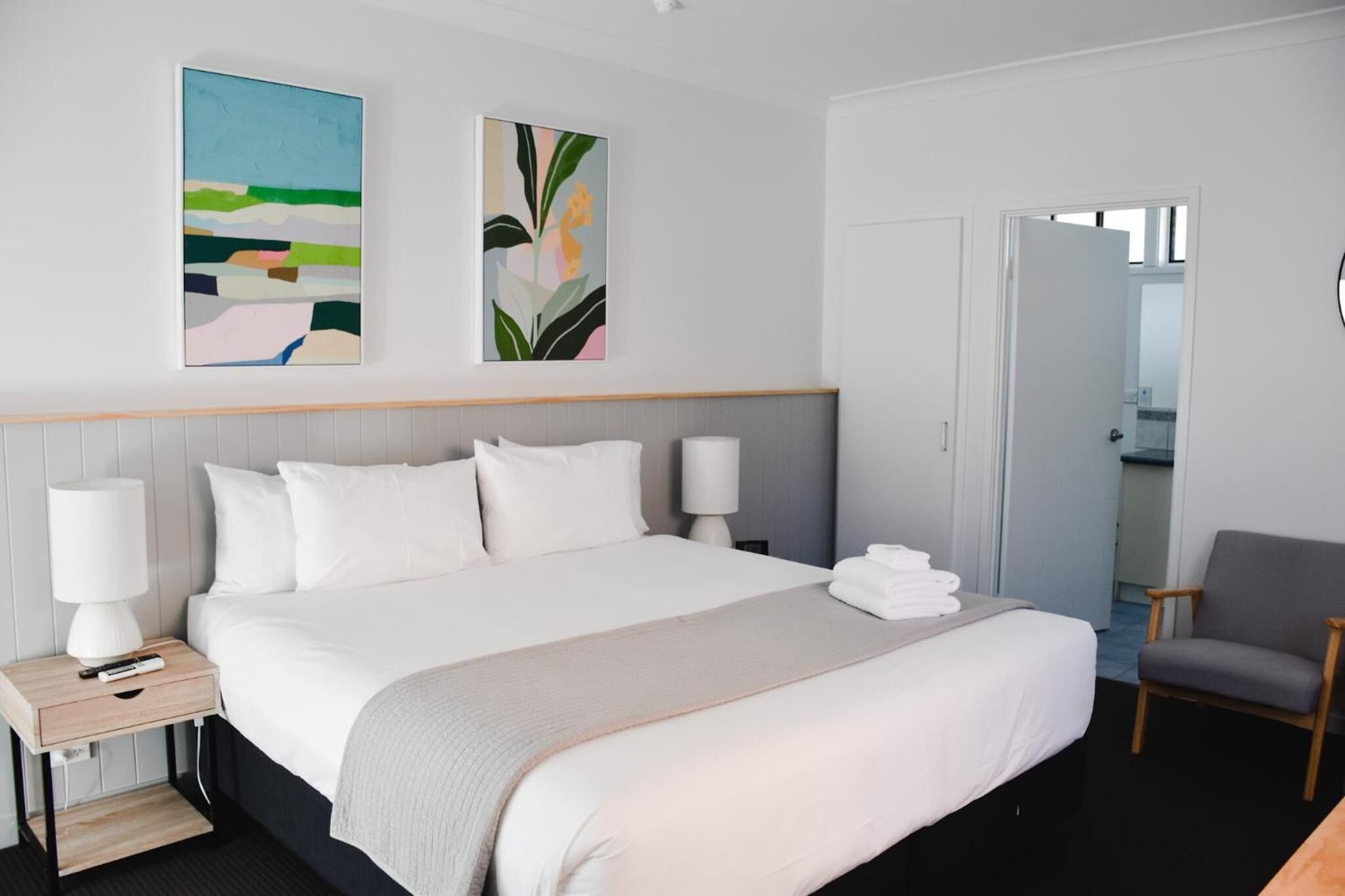 A hotel room with a large bed, a bedside table with a lamp, a chair, and two wall-mounted artworks.