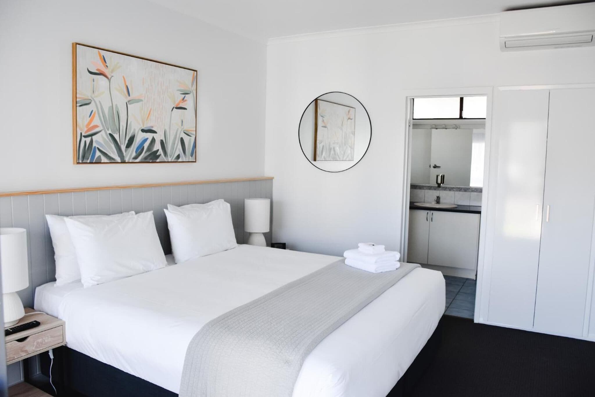 A hotel room with a large bed, a bedside table with a lamp, a wall-mounted artwork, a round mirror, and a view into the bathroom.