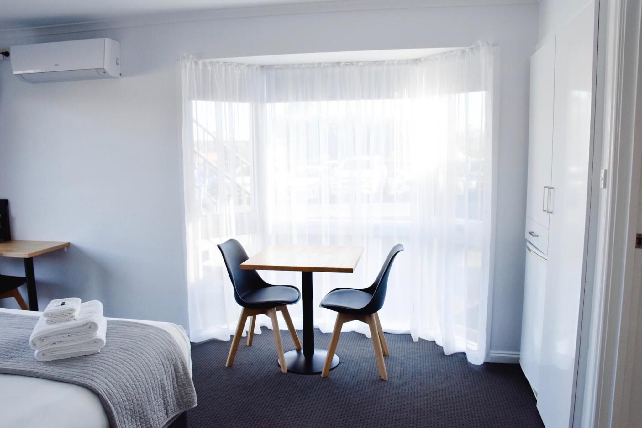 Habitación de hotel con cama, una mesita pequeña con dos sillas, una ventana con cortinas traslúcidas y un armario.