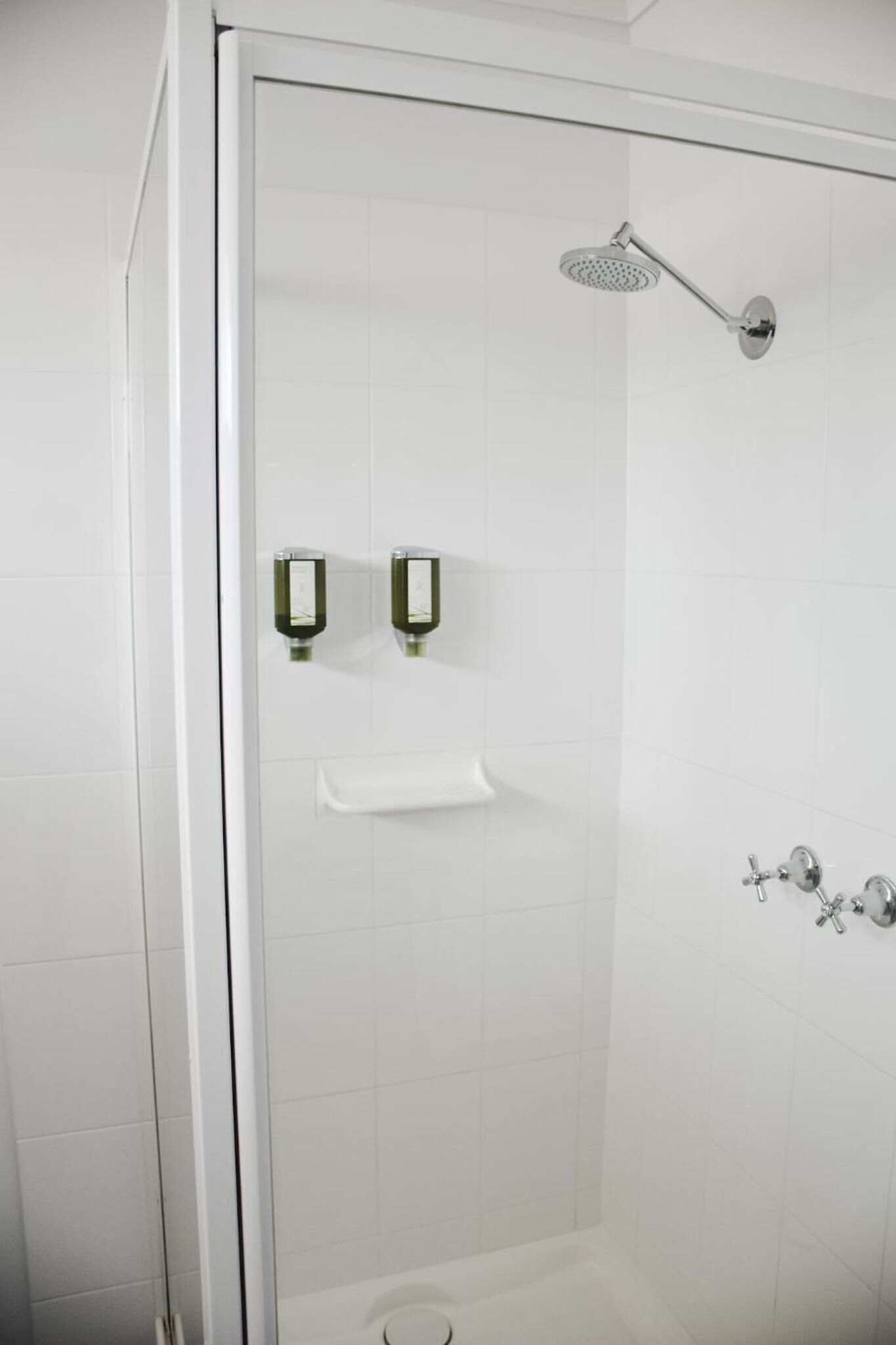 A glass-enclosed shower area with a handheld showerhead, two soap dispensers, and a white towel rack.