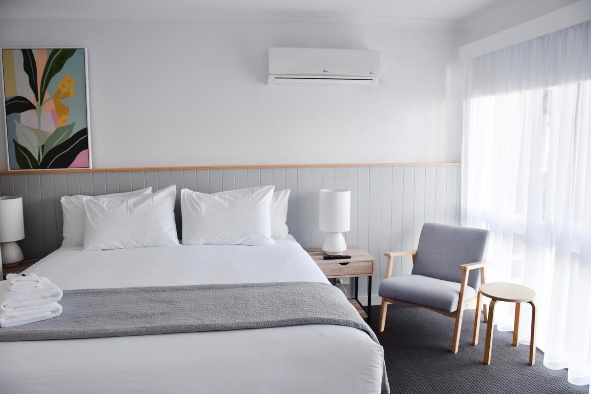 Una camera d'albergo con un letto grande, un comodino con una lampada, una sedia e una finestra con tende leggere.