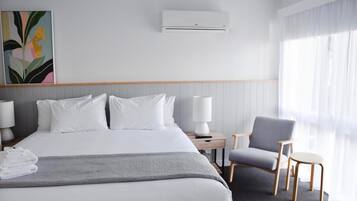 Una camera d'albergo con un letto grande, un comodino con una lampada, una sedia e una finestra con tende leggere.