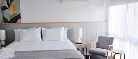 A hotel room with a large bed, a bedside table with a lamp, a chair, and a window with sheer curtains.