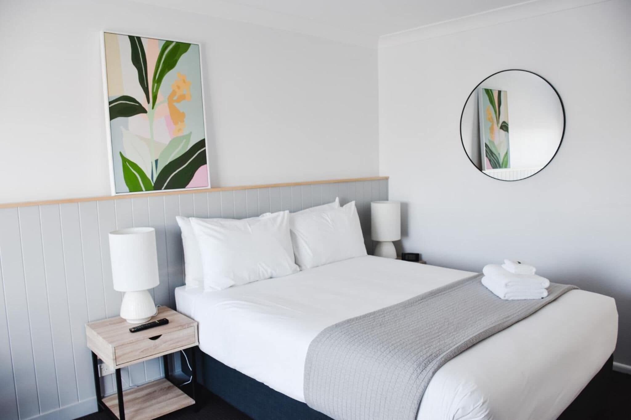 A neatly made bed with white linens, two bedside lamps, a bedside table with a lamp, a wall-mounted mirror, and a framed abstract painting.