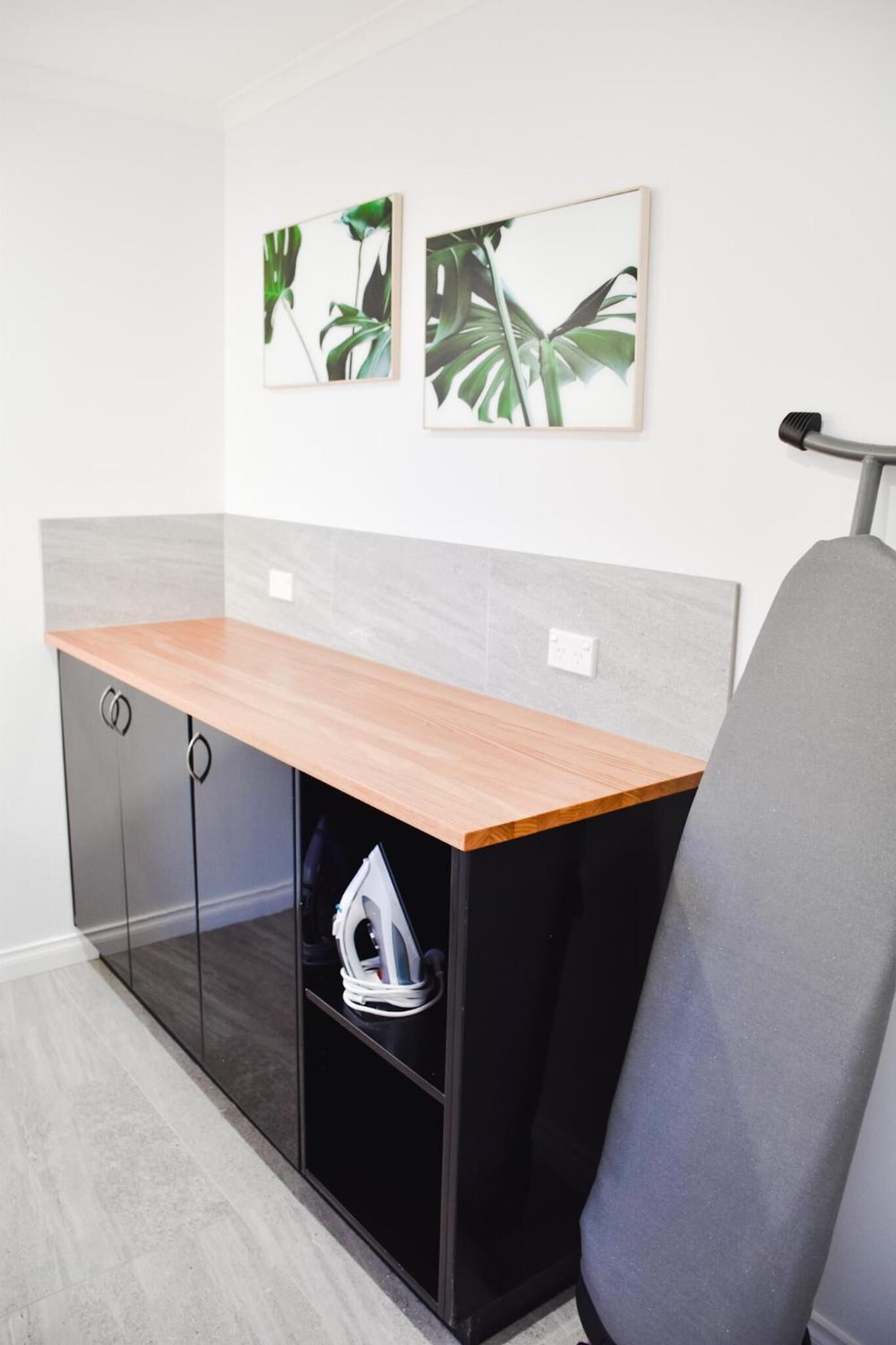 A modern kitchen with a wooden countertop, black cabinets, and a grey chair. There are two framed pictures on the wall.