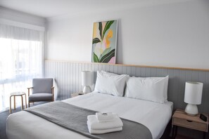 A hotel room with a neatly made bed, a chair, two bedside tables with lamps, and a large window with curtains.