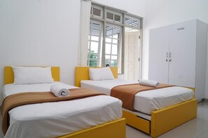 A hotel room with two beds, each with a yellow headboard and white bedding. There is a white wardrobe and a window with blinds. - Omah Sastro (Depok)