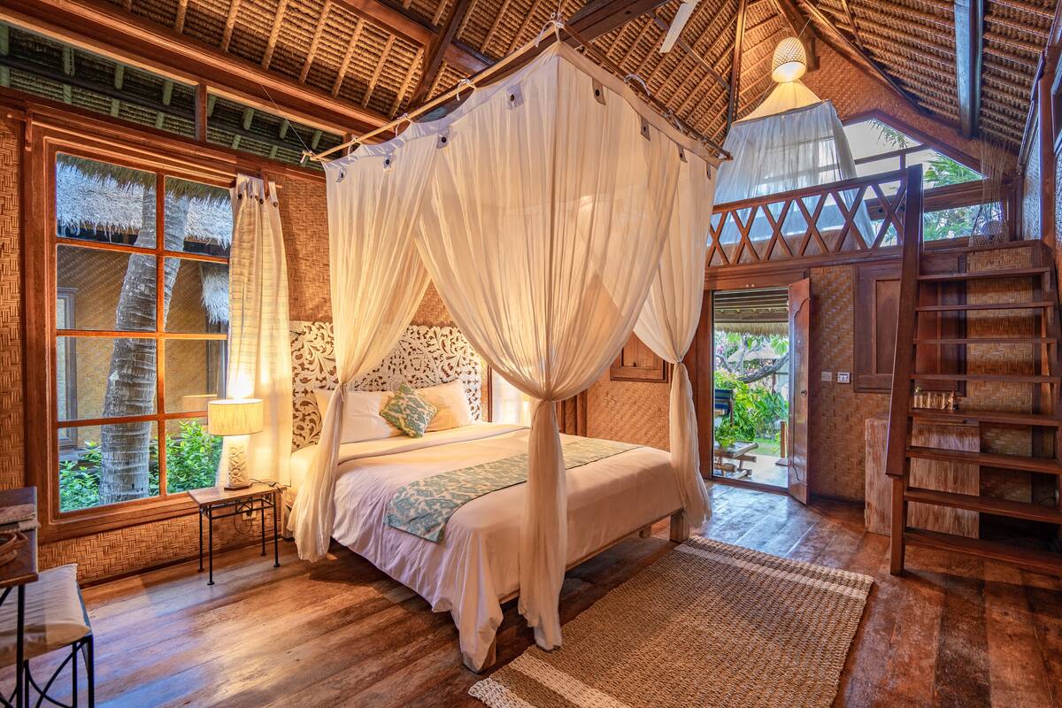 A four-poster bed with a canopy, a wooden floor, a window with a view of greenery, and a wooden ladder.