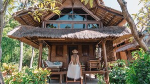 A thatched-roof house with a wooden porch and a view of the surrounding greenery. - Santai Hotel (Karangasem)