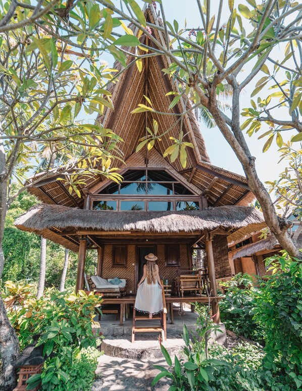 A thatched-roof house with a wooden porch and a view of the surrounding greenery. - Santai Hotel (Karangasem)
