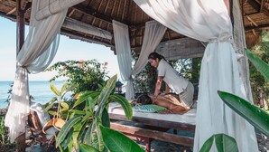 A wooden structure with a thatched roof, a massage table, and a person receiving a massage. - Santai Hotel (Karangasem)