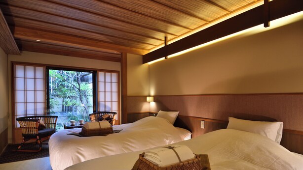 Ein Hotelzimmer mit zwei Betten, einer Holzbalkendecke, Blick auf Grünfläche und einem traditionellen japanischen Stuhl.
