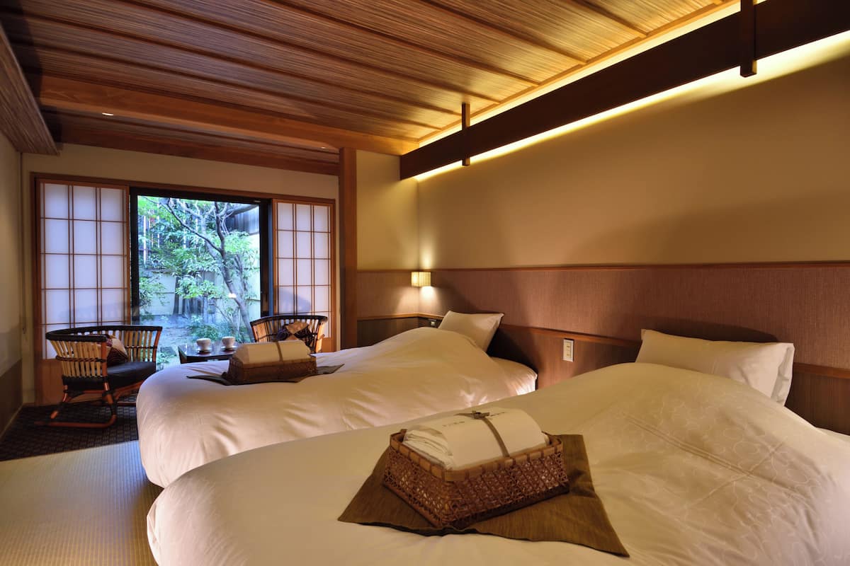 A hotel room with two beds, a wooden ceiling, a view of greenery outside, and a traditional Japanese-style chair.