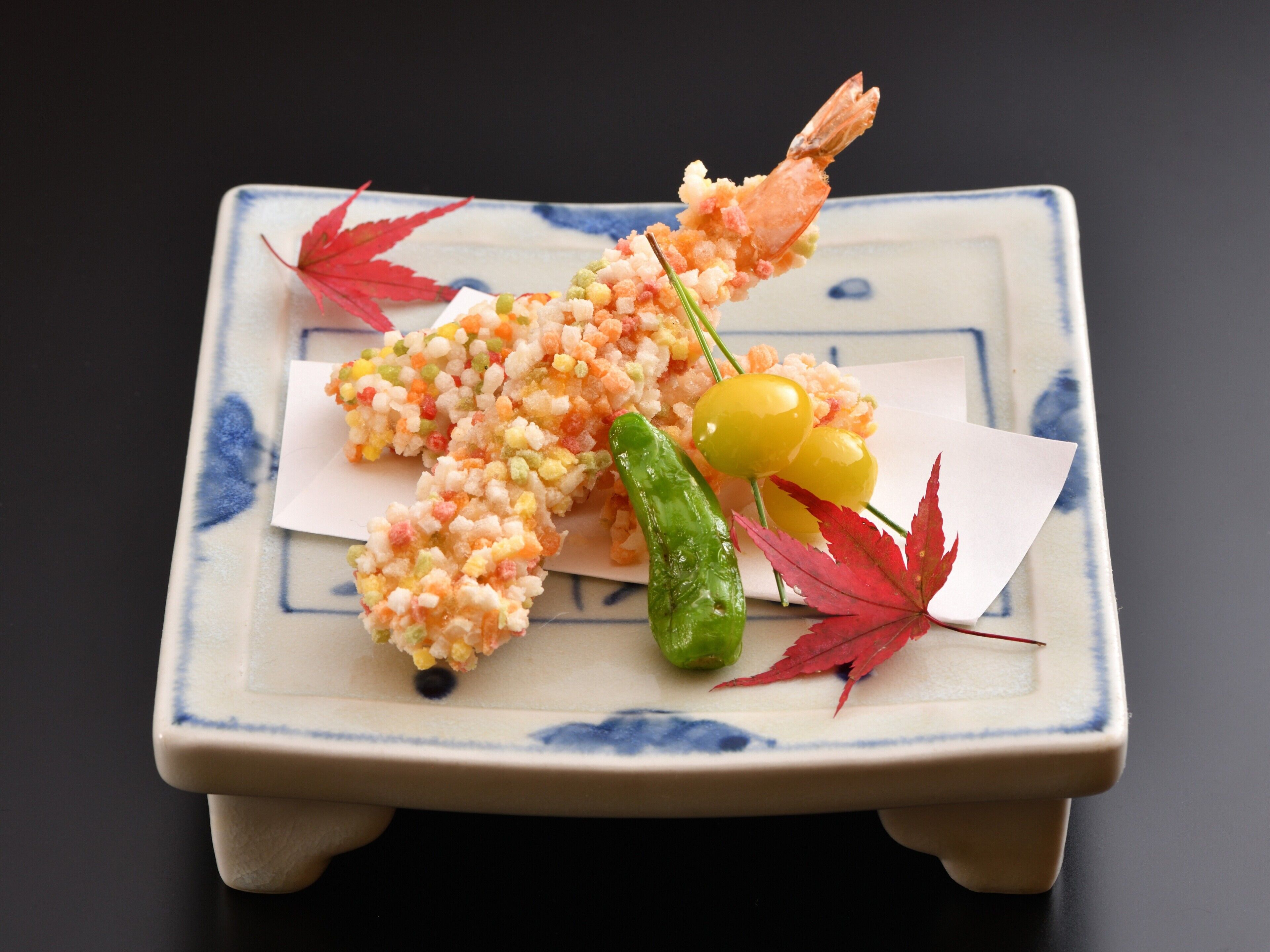 A square ceramic plate with a decorative design, containing a shrimp, cherry tomatoes, and a green chili pepper, with red maple leaves as garnish.