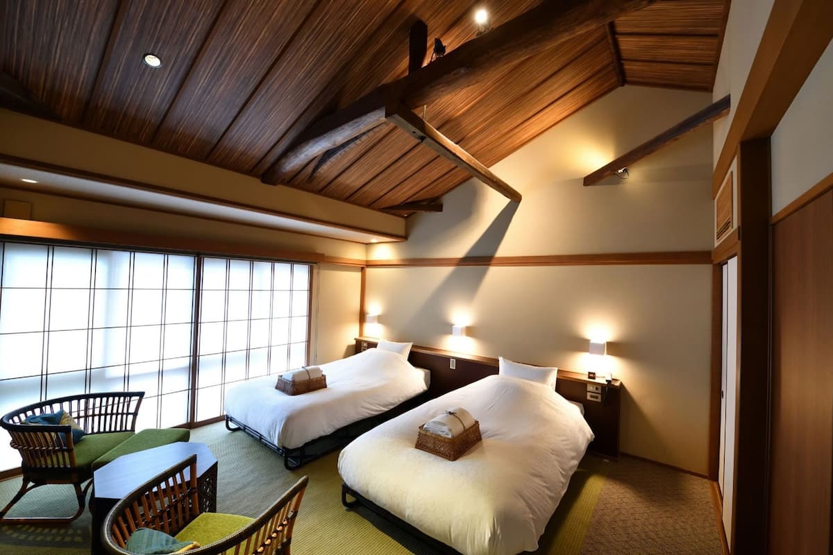 A hotel room with two beds, a dining area with chairs, and a large window.